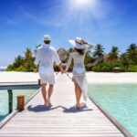 A happy traveler couple in white summer clothing walks down hand in hand a wooden pier towards a tropical paradise island in the Maldives, Indian Ocean